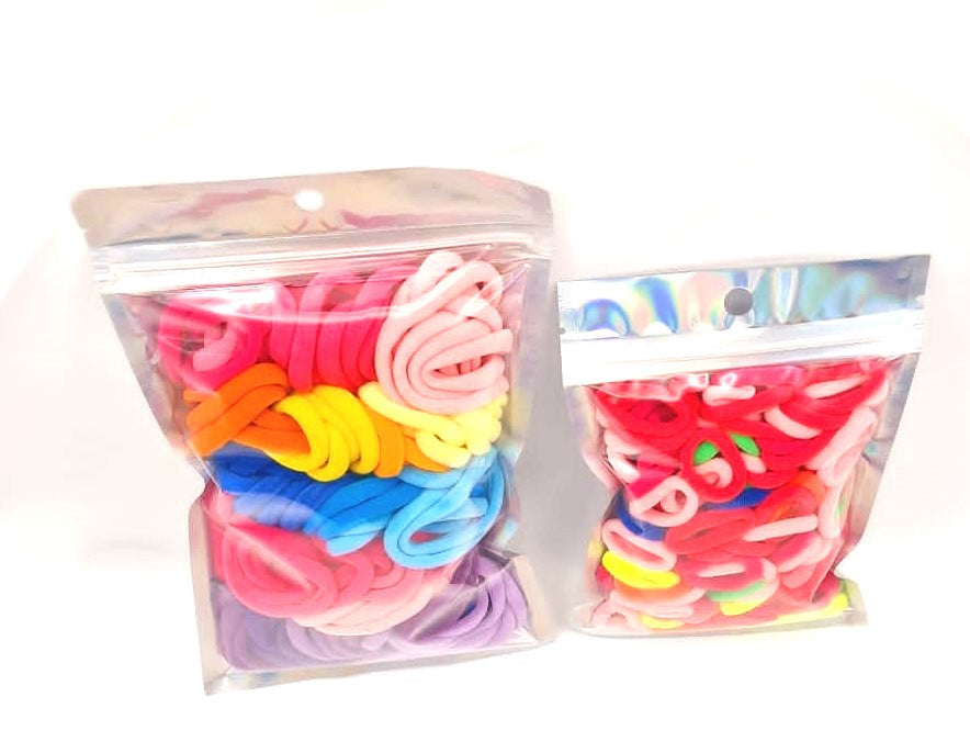 Girls & Baby's Hair Ties Pack of 2: ENAX Multi-Coloured Hair Bands. Two silver packets. First packet to the left consists of (ENAX) Girls Multi-Coloured, medium-sized Hair Ties. Second packet to the right consists of Baby & Toddler's Pink Hair Ties. 80 medium-sized Girls Multi-coloured hair ties and 120 Baby & Toddler's Pink Hair Ties, two packets inside picture. White background. Front Side.
