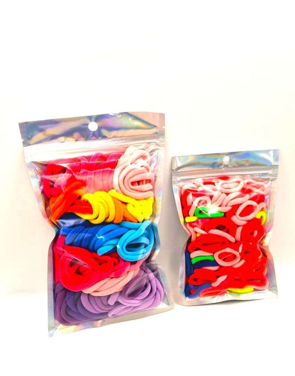 Girls & Baby's Hair Ties Pack of 2: ENAX Multi-Coloured Hair Bands. Two silver packets. First packet to the left consists of (ENAX) Girls Multi-Coloured, medium-sized Hair Ties. Second packet to the right consists of Baby & Toddler's Pink Hair Ties. 80 medium-sized Girls Multi-coloured hair ties and 120 Baby & Toddler's Pink Hair Ties, two packets inside picture. White background. Front Side.