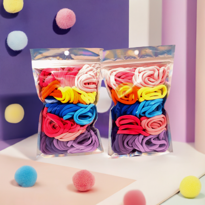 Girls' Hair Ties Pack of 2: ENAX Multi-Coloured Girls Hair Bands, Two silver packet of (ENAX) colorful medium-sized Hair Ties. 80 hair ties inside each pack, two packets in picture. Purple background with purple foam balls. Front Side.