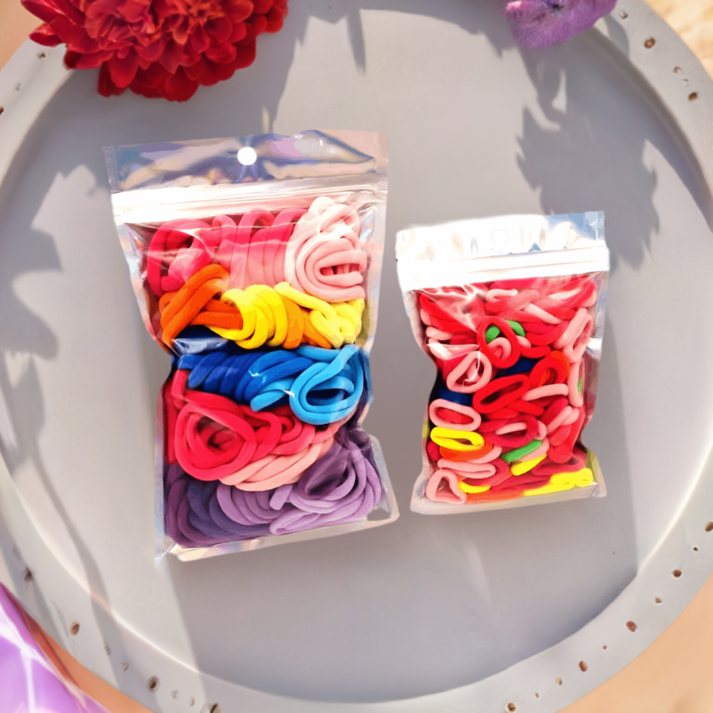 Girls & Baby's Hair Ties Pack of 2: ENAX Multi-Coloured Hair Bands. Two silver packets. First packet to the left consists of (ENAX) Girls Multi-Coloured, medium-sized Hair Ties. Second packet to the right consists of Baby & Toddler's Pink Hair Ties. 80 medium-sized Girls Multi-coloured hair ties and 120 Baby & Toddler's Pink Hair Ties, two packets inside picture. Silver Tray with red flower. Front Side.
