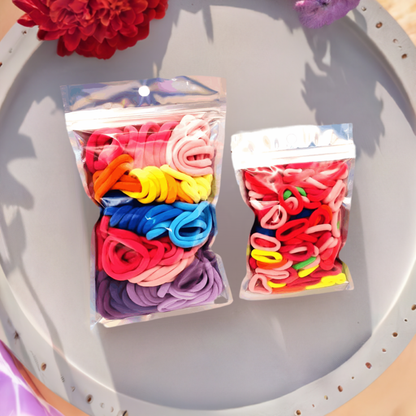 Girls & Baby's Hair Ties Pack of 2: ENAX Multi-Coloured Hair Bands. Two silver packets. First packet to the left consists of (ENAX) Girls Multi-Coloured, medium-sized Hair Ties. Second packet to the right consists of Baby & Toddler's Pink Hair Ties. 80 medium-sized Girls Multi-coloured hair ties and 120 Baby & Toddler's Pink Hair Ties, two packets inside picture. Silver Tray with red flower. Front Side.