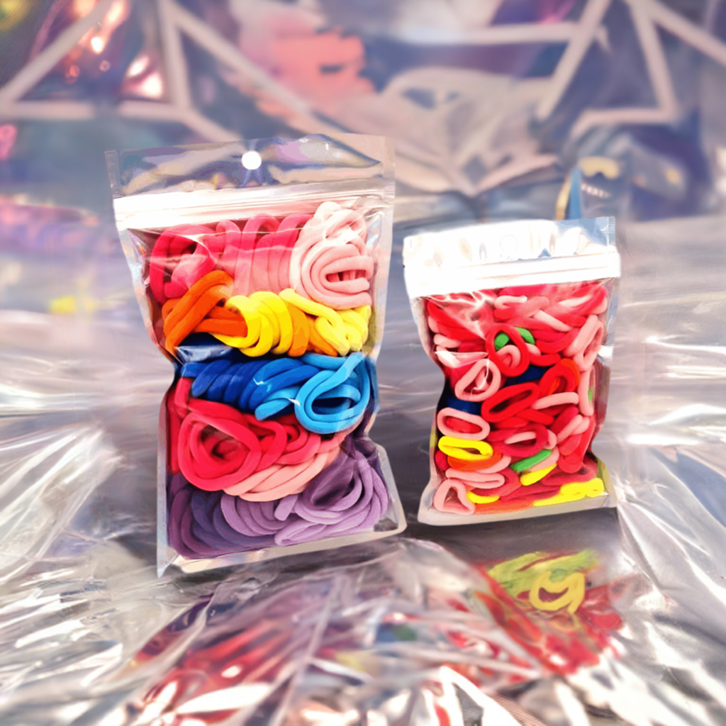 Girls & Baby's Hair Ties Pack of 2: ENAX Multi-Coloured Hair Bands. Two silver packets. First packet to the left consists of (ENAX) Girls Multi-Coloured, medium-sized Hair Ties. Second packet to the right consists of Baby & Toddler's Pink Hair Ties. 80 medium-sized Girls Multi-coloured hair ties and 120 Baby & Toddler's Pink Hair Ties, two packets inside picture. Silver foiled background. Front Side.