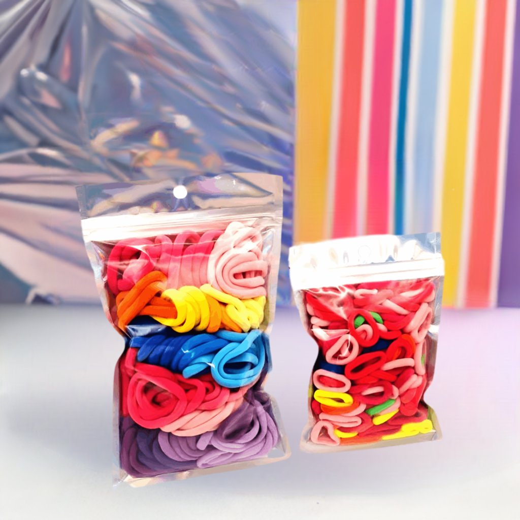 Girls & Baby's Hair Ties Pack of 2: ENAX Multi-Coloured Hair Bands. Two silver packets. First packet to the left consists of (ENAX) Girls Multi-Coloured, medium-sized Hair Ties. Second packet to the right consists of Baby & Toddler's Pink Hair Ties. 80 medium-sized Girls Multi-coloured hair ties and 120 Baby & Toddler's Pink Hair Ties, two packets inside picture. Colourful stripy background. Front Side.